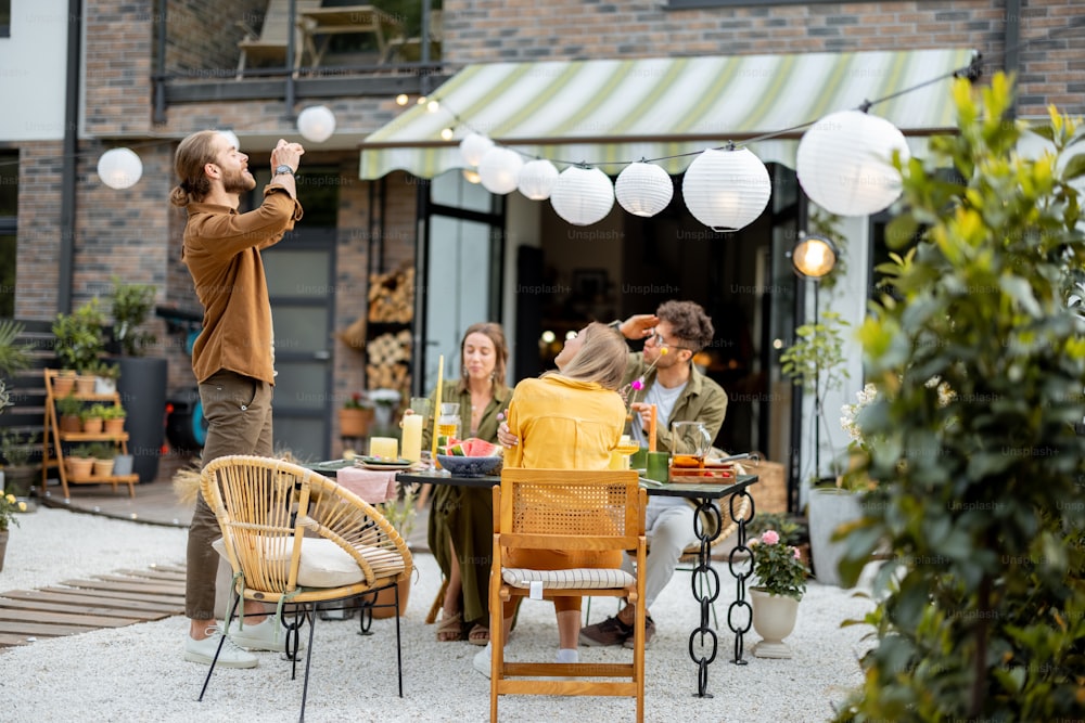 A group of young friends having great summertime, dining and talking at backyard of the country house. Man photographing on phone