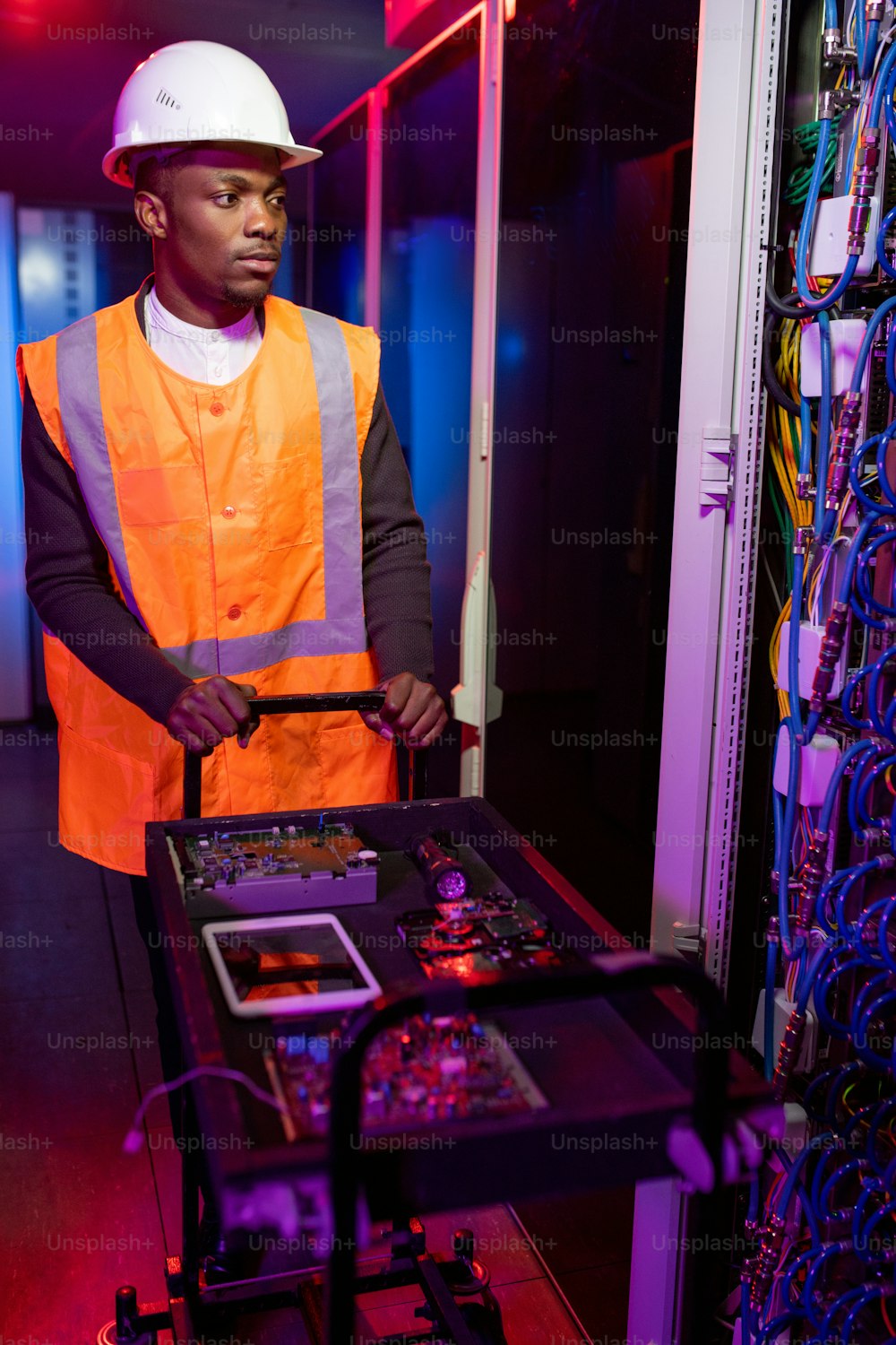 Joven y serio especialista en reparación de servidores negros en cascos y chalecos que empujan el carro con microchips y placas de circuito en el centro de datos en color neón