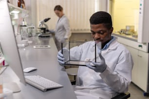 Joven africano en ropa de trabajo mirando al ratón blanco en una caja de vidrio mientras trabaja en el laboratorio