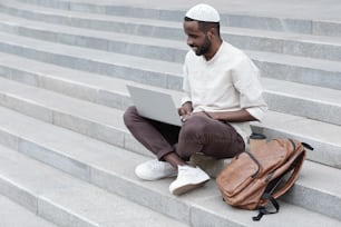 Cheerful handsome young Black man in kufi sitting with laptop between knees and watching webinar outdoors
