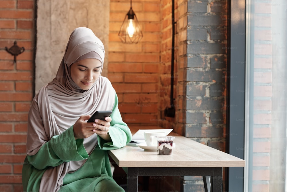 Content young Muslim woman in hijab sitting at high table in loft cafe and texting sms on phone