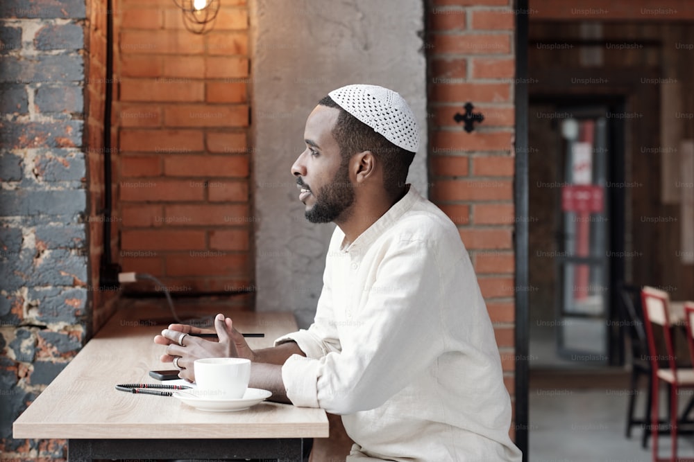 Giovane islamico ispirato pensoso in berretto kufi seduto al tavolo in un caffè loft e guardando fuori dalla finestra
