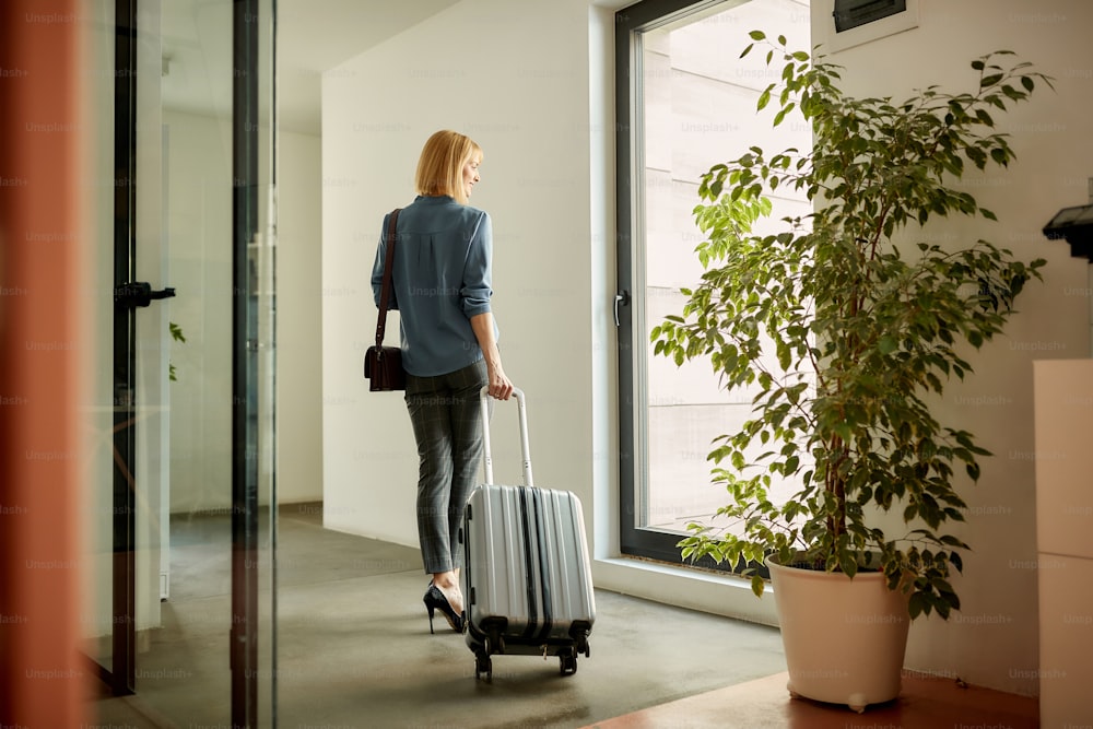 Imprenditrice felice con la valigia che guarda attraverso la finestra in un corridoio mentre va in viaggio d'affari.