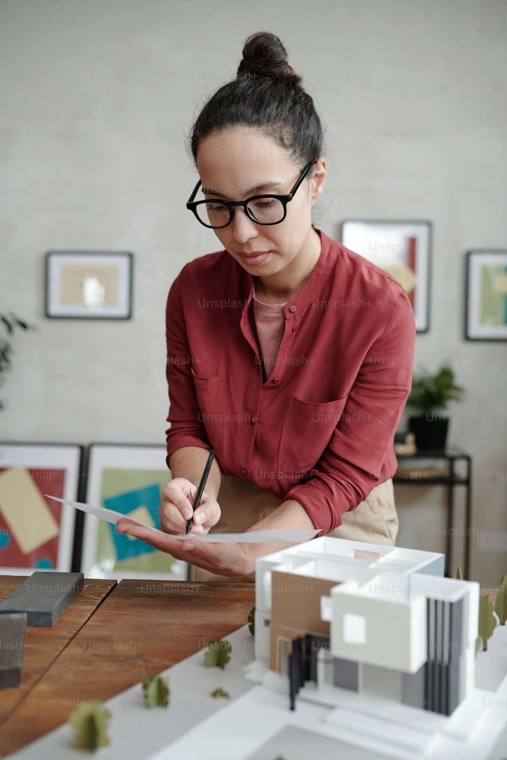 Focused young Hispanic architect in eyeglasses standing at wooden desk with modern model of building and adding information on blueprint