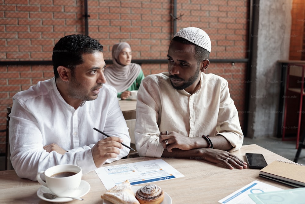 イスラムの衣装を着た忙しい若い多民族のビジネスマンがカフェのテーブルに座り、チャートについて話し合っています