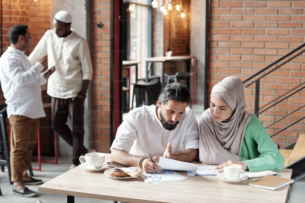 Arabian man with beard pointing at document while discussing statistics with female colleague in cafe