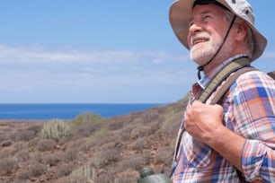 Senior man traveler enjoying outdoors excursion walking between mountain and sea, wearing cap and backpack