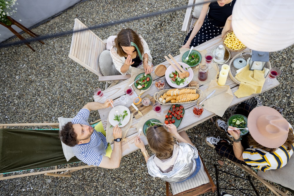 Un gruppo di amici che cenano al tavolo di legno splendidamente decorato pieno di cibo sano, fanno un picnic su una terrazza sul tetto. Vista dall'alto