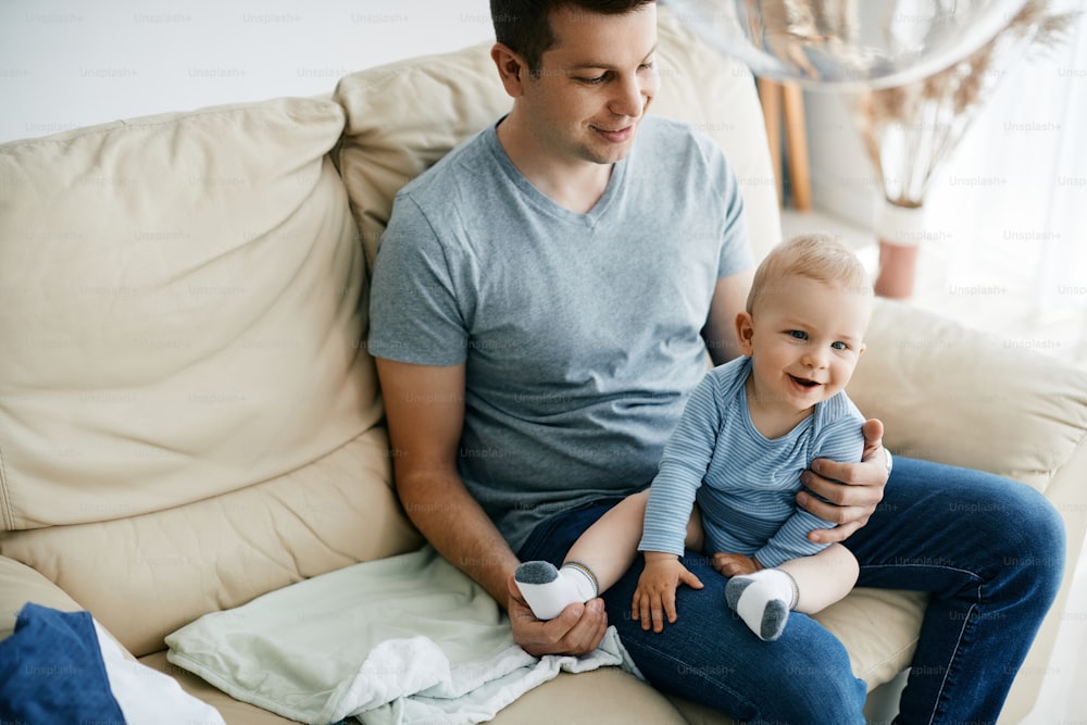 Cute baby boy and his father enjoying together while relaxing on he sofa in the living room.