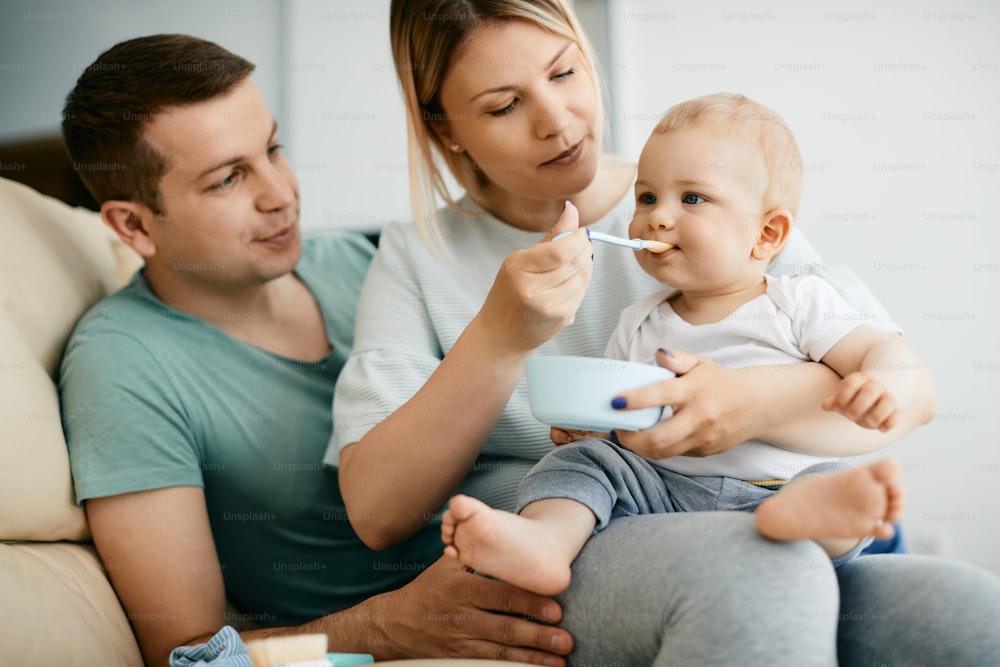 Cute little boy eating baby food while being fed by his parents at home.