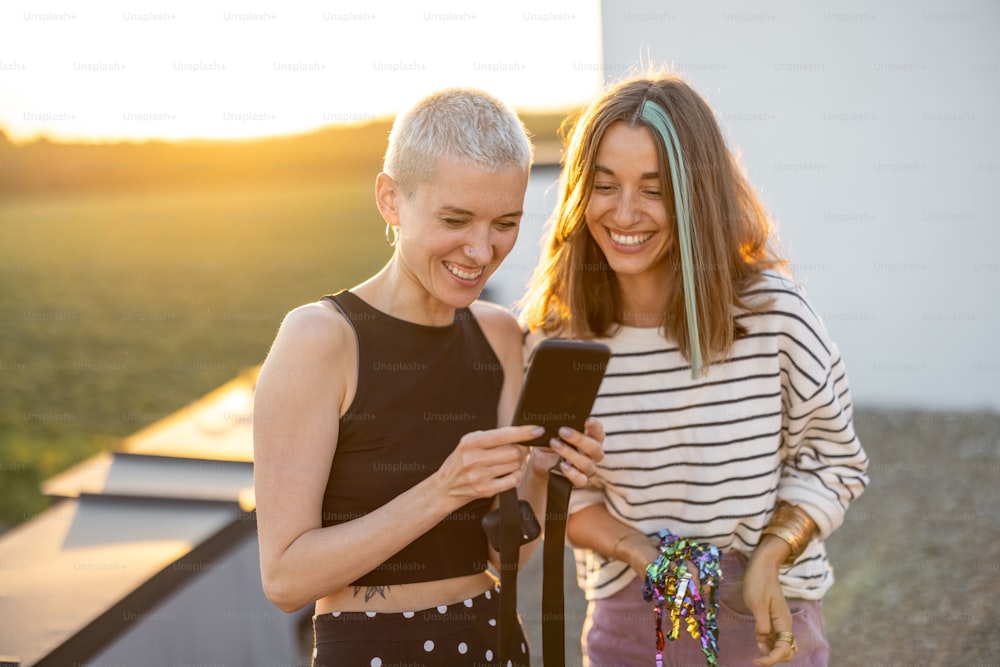 Dois amigos elegantes se divertem, mostrando algo no telefone e rindo ao ar livre. Hipsters e millennials saem no terraço da cobertura ao pôr do sol