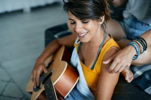 Giovane donna felice che suona acustica per la sua ragazza mentre si rilassa insieme a casa.