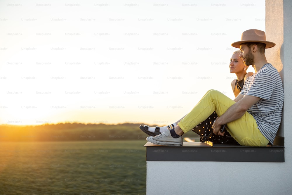La coppia elegante ha una conversazione ravvicinata mentre è seduta insieme sul tetto con una splendida vista sul tramonto sulla natura. Stile di vita spensierato e concetto di romanticismo