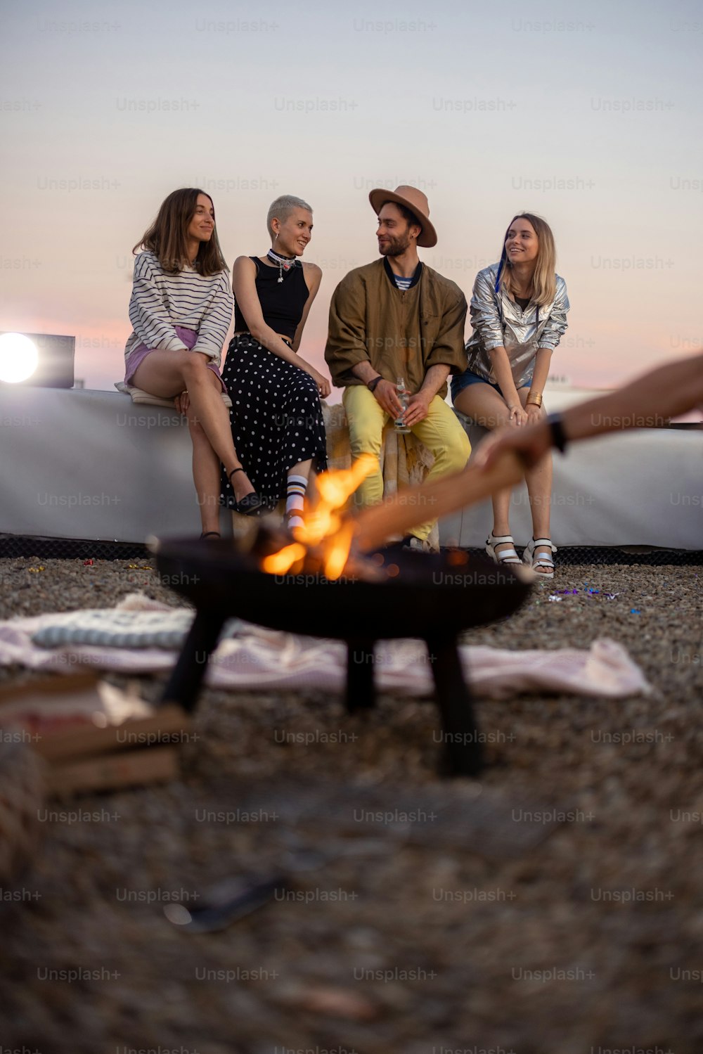 Group of young stylish friends sitting near the fireplace, hanging out together on the rooftop terrace at dusk. Enjoy summertime and beautiful sunset view