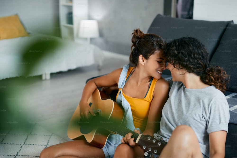 Feliz pareja femenina mostrando afecto mientras una de ellas toca la guitarra acústica en casa.