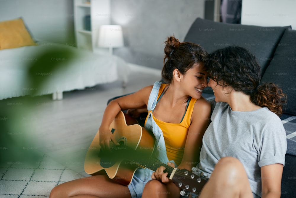 Happy female couple showing affection while one of them is playing acoustic guitar at home.