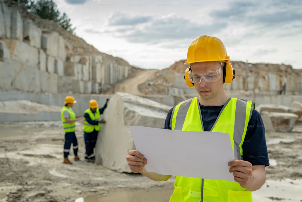 Young serious foreman in protective workwear looking at sketch on blueprint