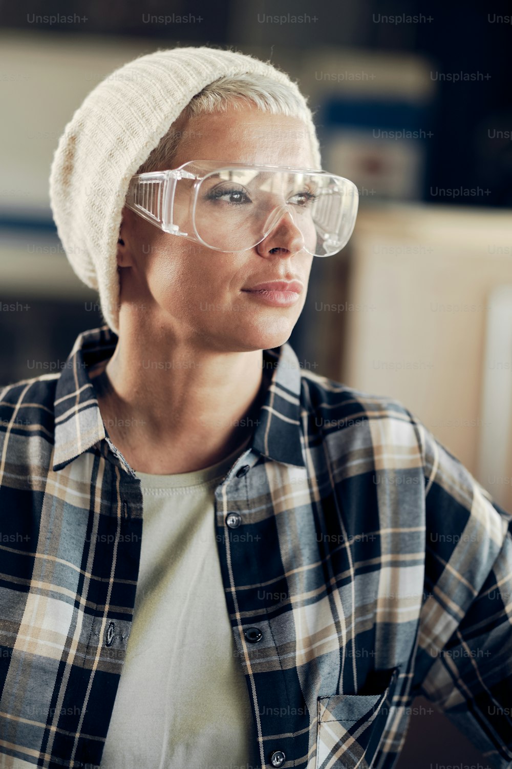 Retrato de perto da mulher em seus 40 anos trabalhando na carpintaria, usando chapéu, óculos de proteção e camisa xadrez