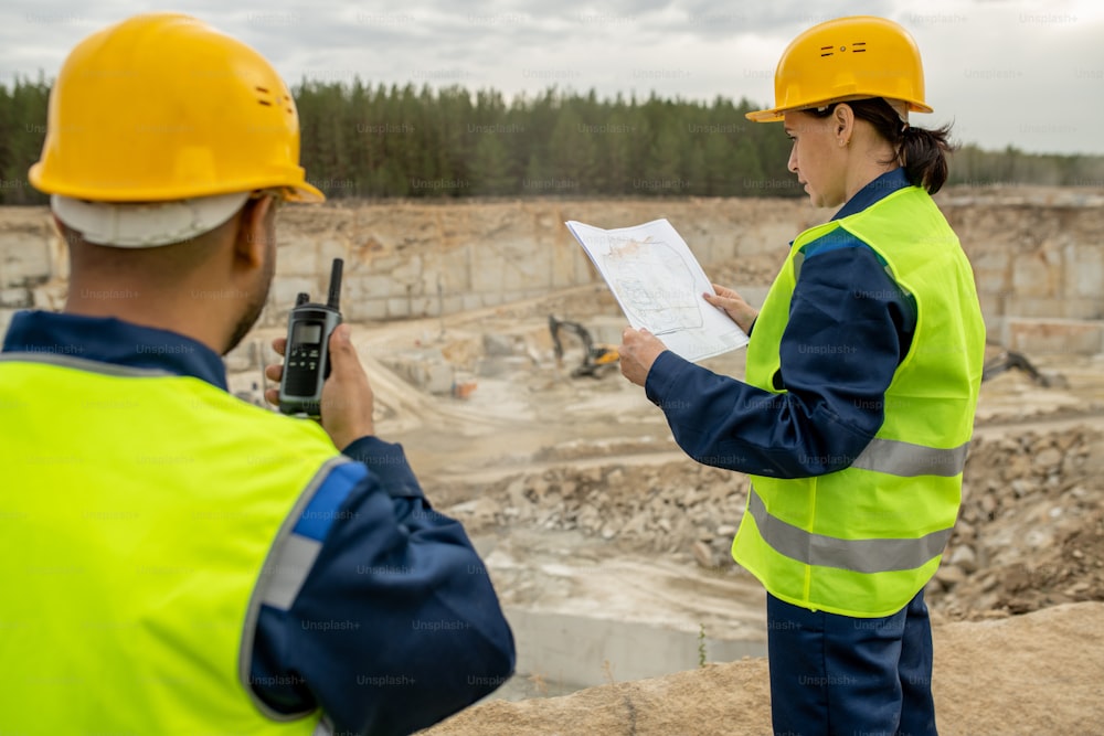 Female engineer looking at sketch and her colleague using walkie-talkie at construction site