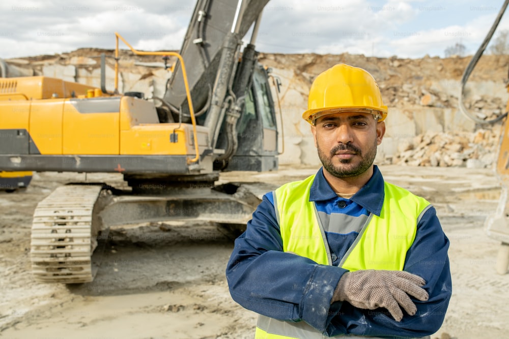 Constructor serio en ropa de trabajo cruzando los brazos en el pecho contra la excavadora