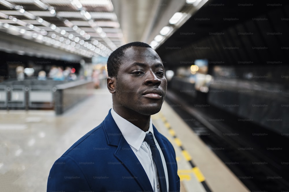 Retrato de un hombre negro confiado sonriente que viste traje azul y corbata en la estación de metro, está mirando el túnel, esperando que llegue el tren