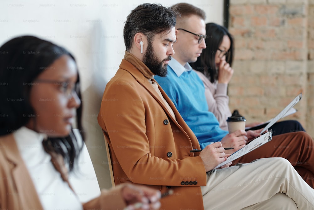 Group of serious young intercultural people sitting in row against wall