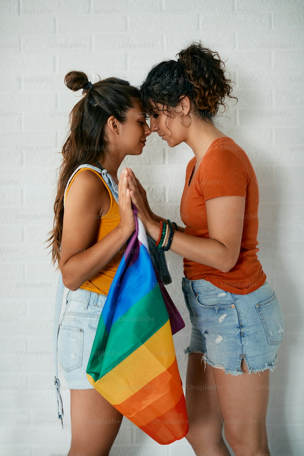 Young homosexual women touching and noses while standing face to face and holding rainbow flag.
