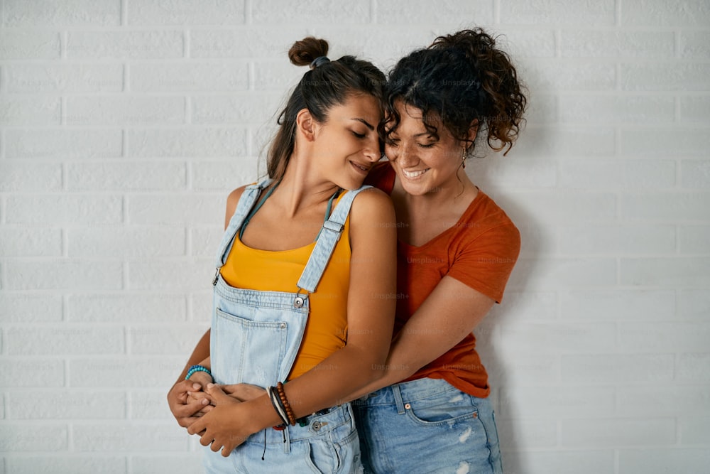 Happy female couple standing embraced against the wall.