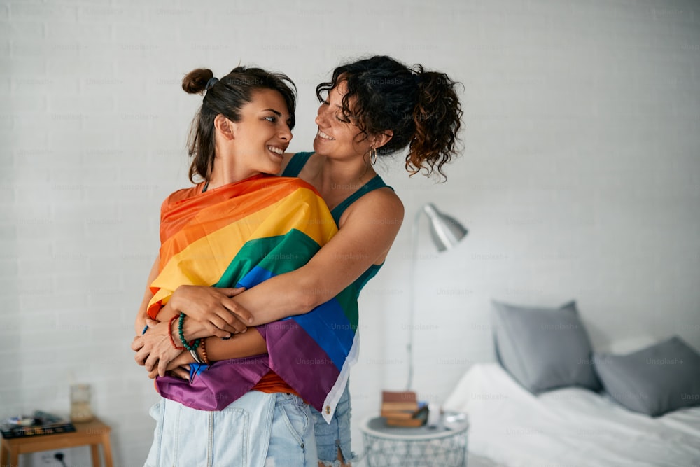 Giovane coppia lesbica innamorata che si abbraccia e si guarda mentre è avvolta nella bandiera arcobaleno.