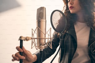Portrait of beautiful curly woman in leather jacket recording vocals in music studio on professional sound equipment