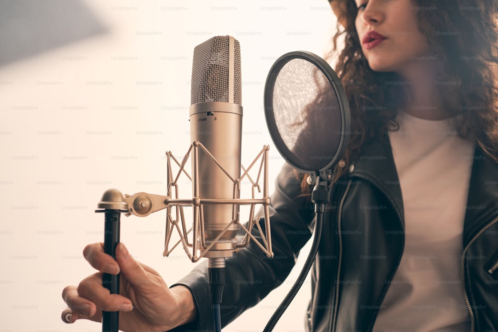 Ritratto di bella donna riccia in giacca di pelle che registra la voce in studio musicale su apparecchiature audio professionali