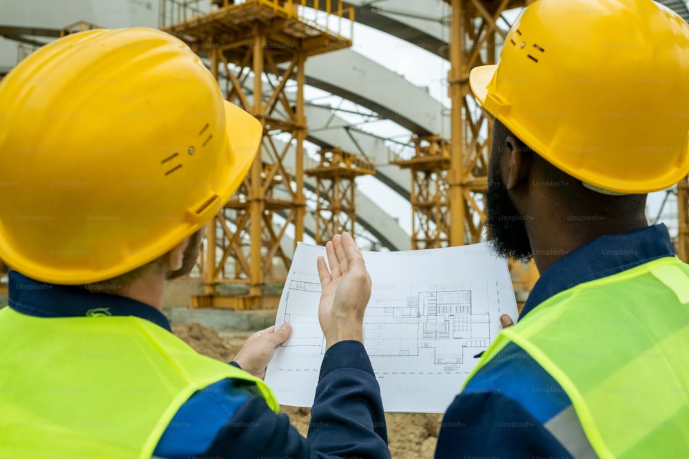 Rear view of two workers in work helmets comparing the blueprint with the building under construction while standing outdoors