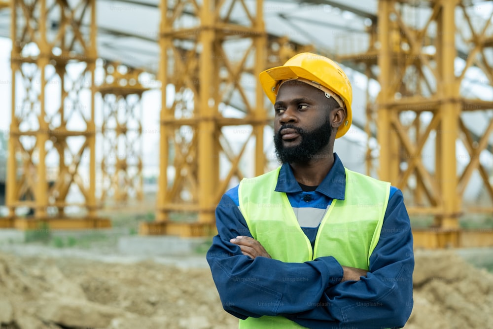 Trabajador de la construcción africano con casco de trabajo de pie con los brazos cruzados en el sitio de construcción y mirando hacia otro lado