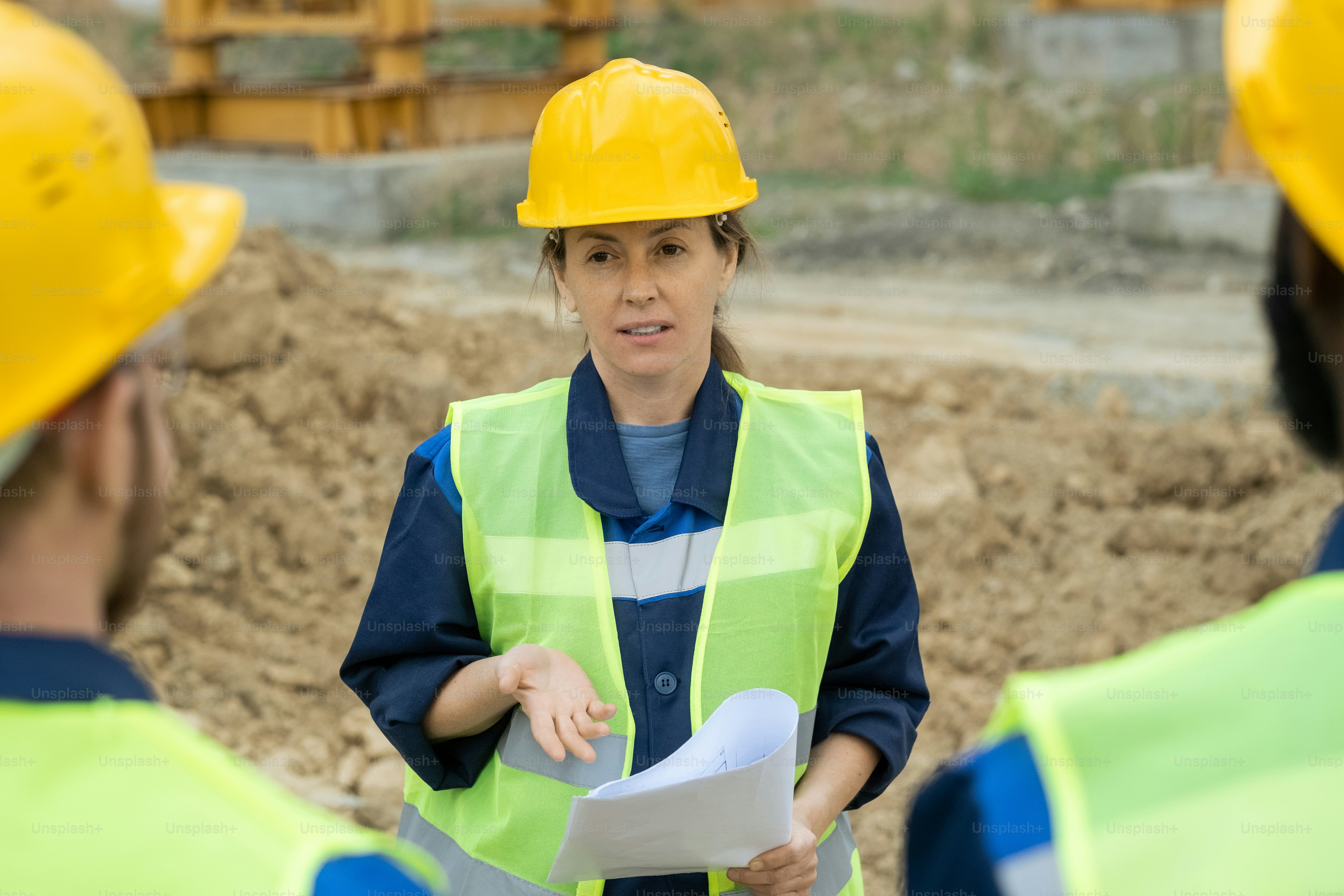 Foreman in work helmet discussing blueprint with workers during construction site outdoors