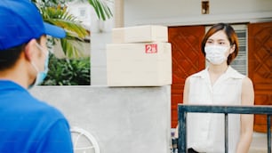 El joven mensajero de entrega postal usa una máscara facial que maneja la caja de paquetes para enviar al cliente en casa y la mujer asiática recibe el paquete entregado al aire libre. Estilo de vida, nueva normalidad después del concepto de coronavirus.