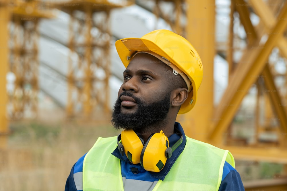 Constructor africano con auriculares y en casco de trabajo mirando la construcción mientras trabaja en el sitio