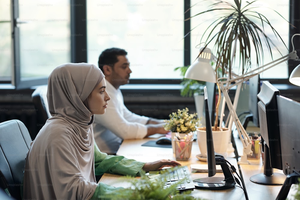 Young Muslim businesswoman in hijab and her male colleague computing by workplace