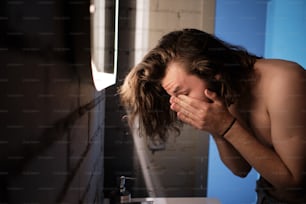 Portrait d’un jeune homme aux cheveux longs se lavant le visage, les mains sur le visage, dans l’évier