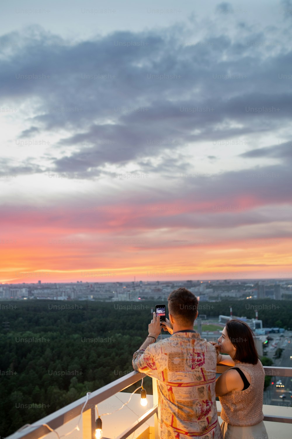Young couple standing on rooftop patio while photographing colorful cloudy sky at sunset