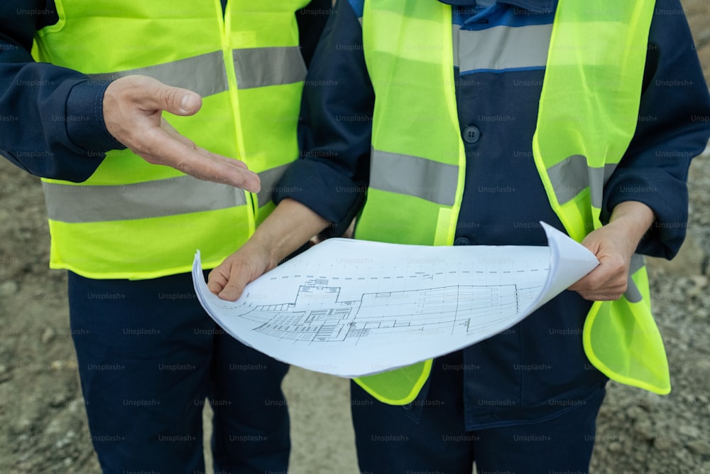 Close-up of two engineers in reflective clothing examining blueprint of new building outdoors