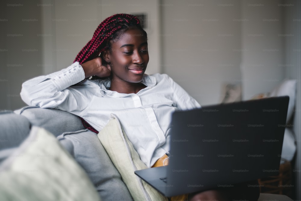 The portrait a dainty young black female with red braids is sitting on a sofa in a living room while working at home and using her laptop to have an online business meeting by a group video call