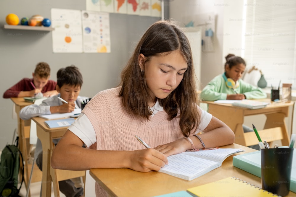 Caucasian elementary schoolgirl making notes while sitting by desk at lesson