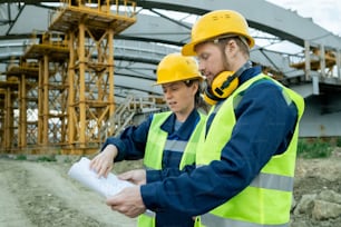 Two workers in work helmets examining blueprint during their work on construction site