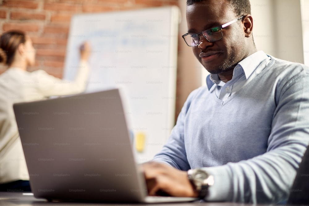 Hombre de negocios negro sonriente trabajando en una computadora mientras su colega escribe en una pizarra en el fondo.
