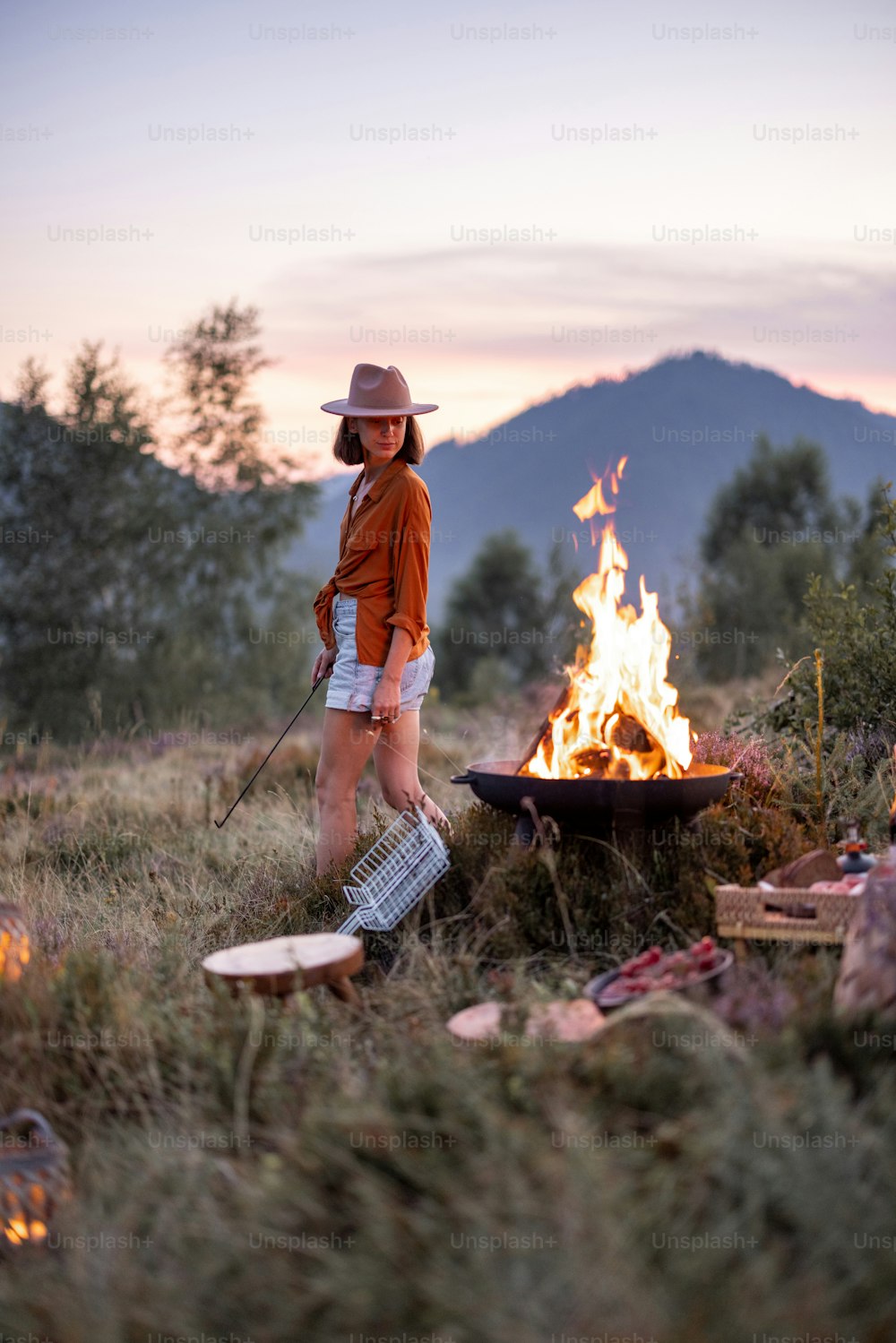 Stilvolle Frau mit einem schönen Picknick mit Lagerfeuer und herrlichem Blick auf die Berge am Abend. Naturgenuss und Reisen in den Bergen Konzept