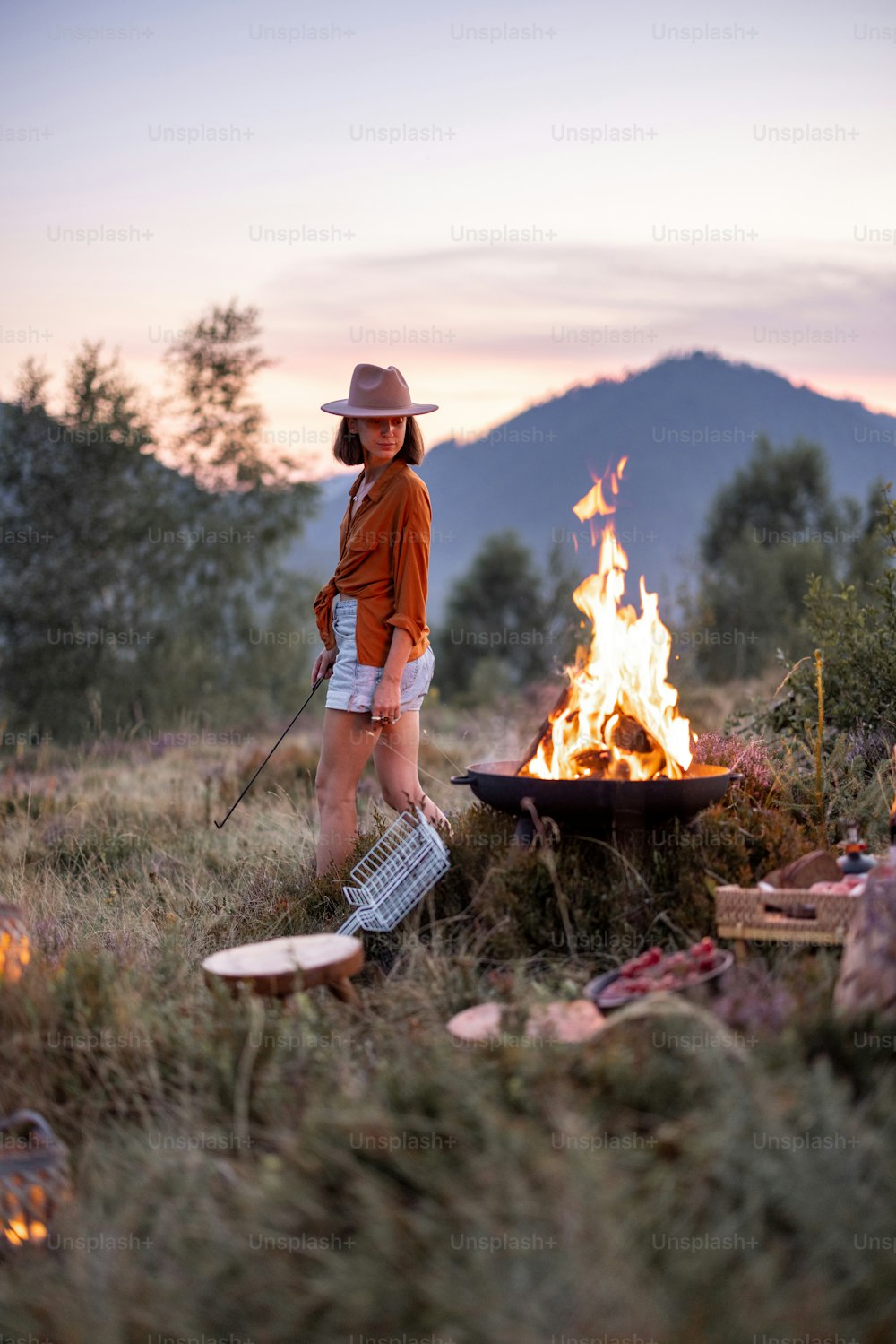 Stylish woman having a beautiful picnic with bonfire and great view on the mountains on the evening. Enjoyment of nature and traveling in the mountains concept