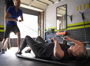 Male personal trainer holds onto elastic rubber resistance bands while an elderly senior man lying on matt on floor and does bicep curl for strength and rehabilitation exercise