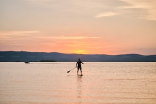 Full body shot of a man with paddle standing on wooden board in quiet water, surfing on sunset with scenic mountain view