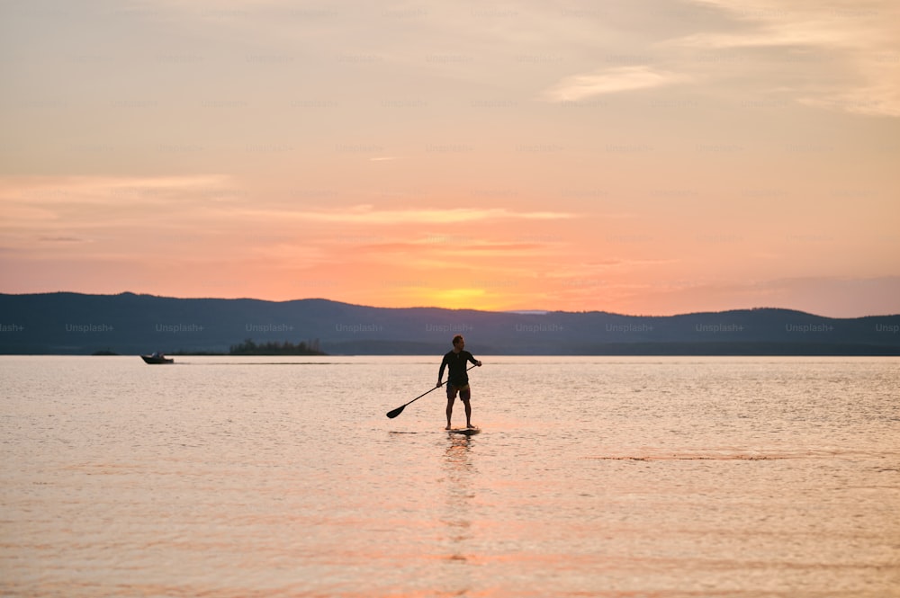 Foto de corpo inteiro de um homem com remo em pé na prancha de madeira em águas tranquilas, surfando no pôr do sol com vista panorâmica da montanha