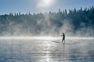 Silhouette eines Mannes, der am frühen Morgen auf dem Surfbrett paddelt, wenn Nebel in den Bergen aufsteigt